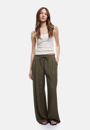 Frau mit langen braunen Haaren, die ein weißes ärmelloses Top, lockere olivgrüne Hose mit Kordelzug und schwarze Sandalen trägt, steht vor weißem Hintergrund.