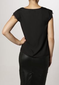 Femme portant un chemisier noir à manches courtes avec bordure en dentelle et une jupe en cuir noire, posant avec une main sur la hanche, dos tourné vers la caméra.