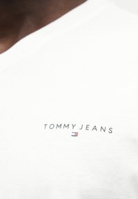 Λευκό βαμβακερό μπλουζάκι με το λογότυπο "TOMMYJEANS" σε μαύρο, με ένα μικρό κόκκινο, λευκό και μπλε διακριτικό από κάτω. Απλός, κλασικός σχεδιασμός.