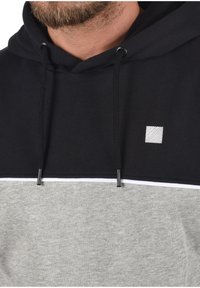Hoodie in zwart en grijs stof, met een witte horizontale streep, koordjes en een klein vierkant logo op de borst.