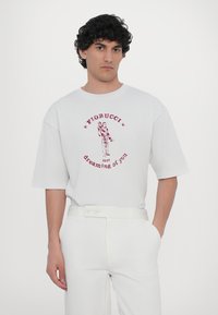 Bijela pamučna majica kratkih rukava s crvenim grafičkim otiskom figure i tekstom "Fiorucci dreaming of you." Glatka tekstura.
