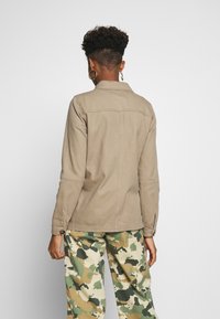 Chaqueta de algodón caqui con mangas largas, cuello y costuras en la parte trasera, combinada con pantalones de patrón de camuflaje. Tonos tierra y diseño casual.