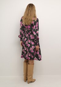 Blumenkleid in Schwarz mit pinken und weißen Orchideenmustern, lange Ärmel mit Manschetten, ausgestellt geschnitten, kombiniert mit beigen knielangen Stiefeln.