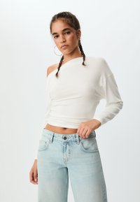 Bijela off-shoulder bluzica dugih rukava s pripijenim dizajnom, uparena s svijetloplavim trapericama visokog struka. Glatka tekstura; svestran stil.