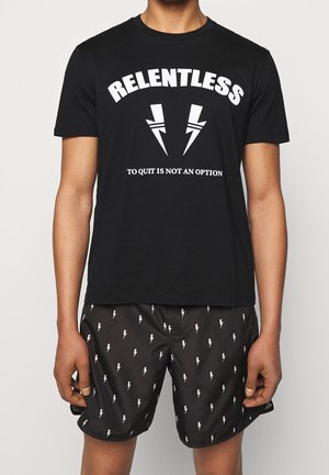 Mężczyzna w czarnej koszulce z napisem "Nieustępliwość, rezygnacja nie jest opcją" oraz w czarnych szortach z białym wzorem błyskawic.