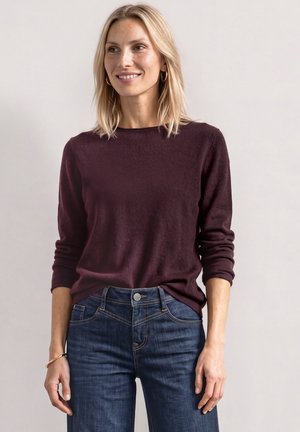 Femme blonde portant des boucles d’oreilles créoles en or, un haut violet foncé à manches longues, un jean bleu et un bracelet fin, debout devant un fond clair uni.
