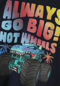 Camiseta navy con un texto en degradado que dice "¡SIEMPRE A LO GRANDE! HOT WHEELS" acompañado de un gráfico de un camión monstruo azul, con grandes neumáticos y acentos coloridos.