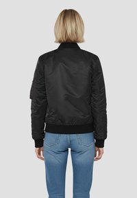 Czarna kurtka bomberka o gładkiej, błyszczącej teksturze, z prążkowanymi mankietami i dołem, z tylną wstawką, bez widocznych oznaczeń. Do kompletu niebieskie dżinsy.