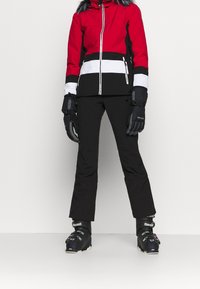 Persona vestida con una chaqueta de esquí roja, negra y blanca, con guantes negros, pantalones de esquí negros y botas de esquí negras, de pie frente a un fondo liso.