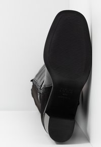 Bottine noire posant le talon contre un mur blanc, montrant la semelle texturée et le talon bloc.