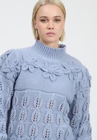 Hellblauer, gestrickter Pullover mit hohem Kragen, der mit floralen Applikationen und einem durchbrochenen Blattmuster verziert ist. Mit strukturiertem und dickem Garn.