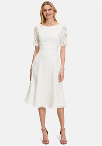 White knee-length dress with a lace bodice, round neckline, and short lace sleeves. Fluid skirt with a soft, flowy texture. Pink heels.