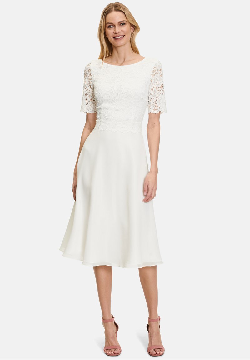 White knee-length dress with a lace bodice, round neckline, and short lace sleeves. Fluid skirt with a soft, flowy texture. Pink heels.