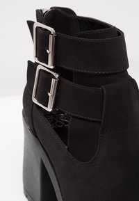 Bottine en daim noir avec deux brides à boucle argentée et un talon haut épais, dotée d'un design découpé sur le côté.