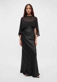 Černé šaty délky po zem z hladké látky s průsvitným vrstveným pláštěm na rukávu. Jednoduchý střih a minimalistický design zdůrazňující elegantní strukturu.
