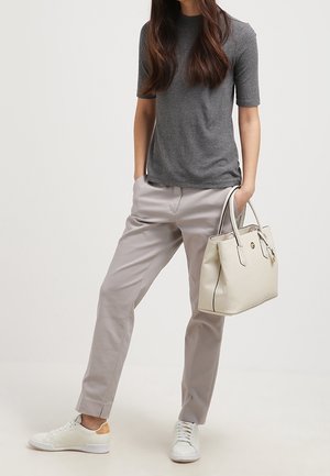Femme portant un haut gris à manches courtes, pantalon gris clair, baskets blanches, tenant un sac à main beige, une main dans la poche, debout devant un fond blanc.