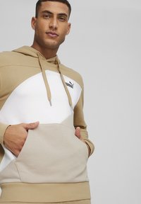 Beżowa i biała bluza z kapturem z kieszenią kangurzą, z białym geometrycznym wzorem i czarnym logo na piersi. Materiał bawełniany.