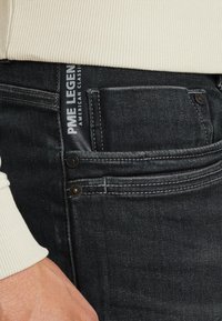 Detailný záber na tmavé džínsy s prednou kapsou a viditeľnou štítkou značky PME Legend, nosené s krémovým rebrovaným rukávom svetra a rukou odpočívajúcou blízko.