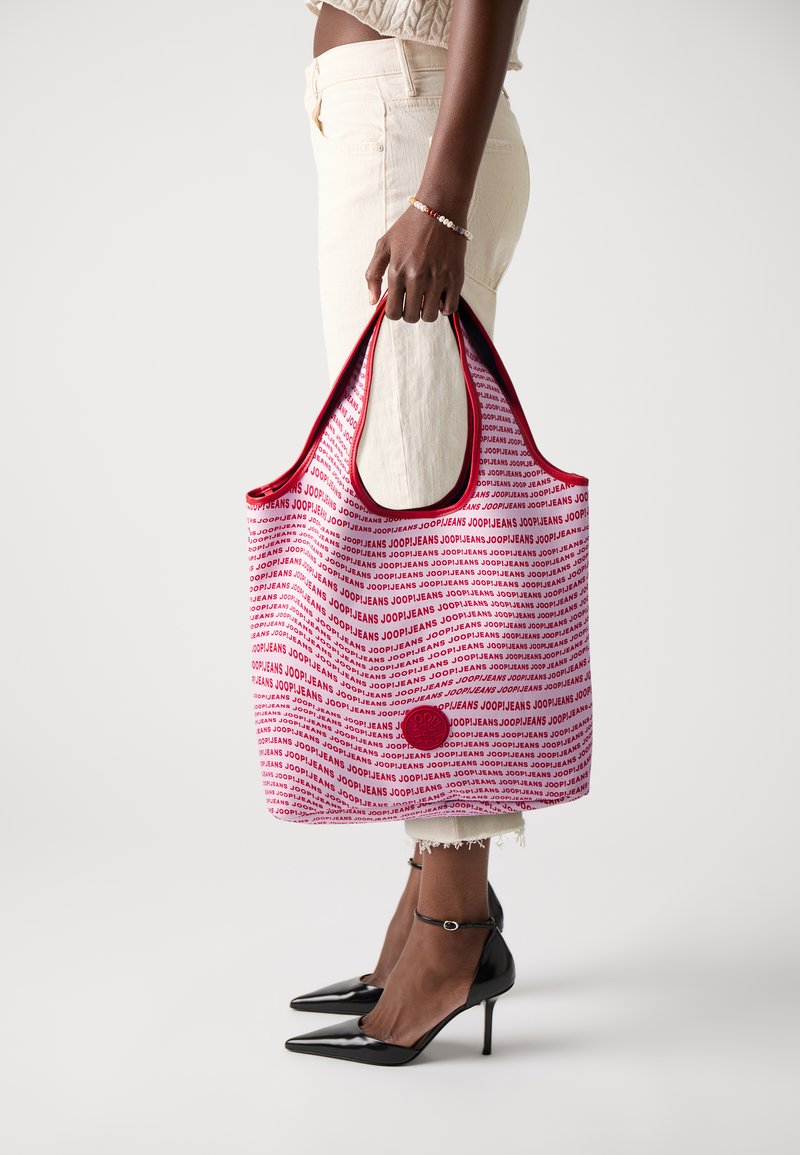 Tote torba v rdeči in beli barvi s potiskanimi besedami "JOOPJEANS." Ima trdno zasnovo, mehak material in okroglo logotipsko podrobnost na sprednji strani.