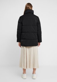 Abrigo acolchado negro con cuello alto y detalle de costura horizontal, combinado con una falda plisada beige y zapatillas de color claro.