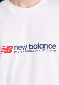 Camiseta de algodón blanca con un destacado logo en rojo y azul de "New Balance" y el texto "Equipo Atlético Profesional". Textura suave.