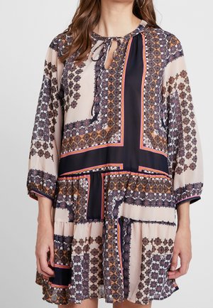 Femme portant une robe à manches longues, longueur au genou, avec des motifs floraux géométriques en beige, noir, orange et rose, dotée d'un col à nouer.