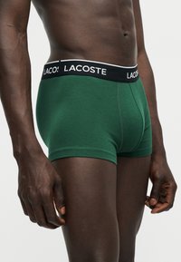Homme portant un boxer vert Lacoste avec une ceinture noire, debout devant un fond clair uni, montrant le torse et le haut des jambes.
