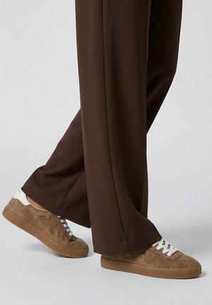 Zapatillas de ante marrón con cordones blancos y suelas de goma, usadas con pantalones sueltos de color marrón oscuro sobre un fondo liso.