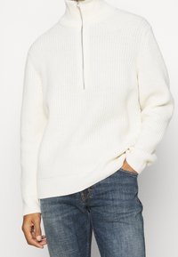 Pull en tricot côtelé blanc avec un col montant et une demi-fermeture éclair à l'avant. Caractérisé par des manches longues et une coupe décontractée, associé à un jean bleu.