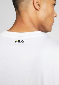 Biała bawełniana koszulka z okrągłym dekoltem. Posiada czarno-zielone logo Fila na plecach w pobliżu dekoltu. Gładka tekstura.