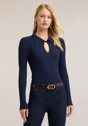 Giovane donna con lunghi capelli biondi che indossa una maglia a coste blu navy a maniche lunghe con colletto e dettaglio ad occhiello, jeans scuri e cintura marrone.