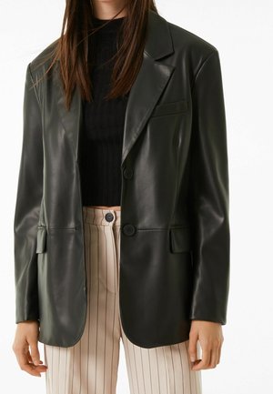 Vrouw draagt een zwarte blazer van imitatieleer over een zwart geribd cropped topje en een beige broek met hoge taille, dunne verticale strepen en een knoopsluiting aan de voorkant.