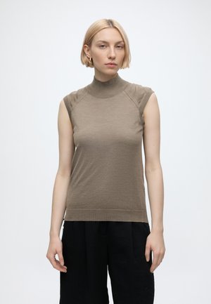 Jeune femme aux cheveux blonds courts portant un haut marron à col roulé sans manches et un pantalon noir sur un fond blanc uni.