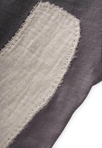 Tela gris con un parche de beige texturizado. El parche tiene bordes sin terminar y costuras prominentes, creando un contraste en color y textura.