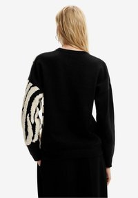 Blond osoba widziana z tyłu, ubrana w czarny sweter z teksturowanym białym i czarnym wzorem w paski przypominającym tygrysa na lewym rękawie.