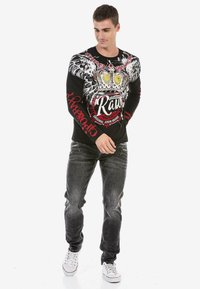 Schwarzes Langarmshirt mit Flügelgrafik, Kronendesign und roten Text. Dazu graue Jeans und weiße Sneaker. Lässiger Schnitt; Baumwollmaterial.