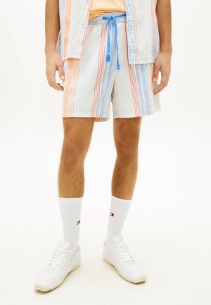 Hombre con pantalones cortos de rayas multicolores con cordón azul, calcetines blancos con pequeño logo de bandera y zapatillas blancas, de pie sobre fondo blanco.