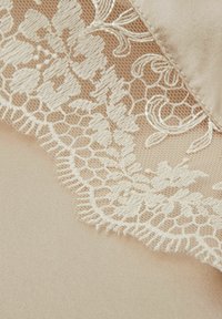 Béžová tkanina s jemným krémovým krajkovým vzorem zdobeným květinovými motivy a zvlněnými okraji, kontrastující s hladkým saténovým povrchem pod ním.