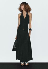 Woman with curly hair wearing a sleeveless black buttoned vest and long black skirt, holding a black woven bag, standing against a white wall.