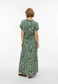 Femme aux cheveux courts portant une robe maxi à manches courtes à motif abstrait vert et blanc, nouée à l'arrière, debout sur un fond blanc.