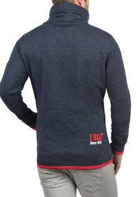 Sudadera de cuello alto en azul marino. Presenta acentos rojos en los puños y el dobladillo, y un logo en la esquina inferior izquierda de la parte trasera.