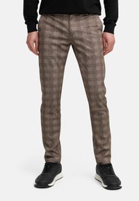 Persona vestida con pantalones marrones a cuadros y zapatillas negras, de pie recta, con las manos relajadas a los lados, contra un fondo blanco liso.