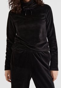 Suéter de terciopelo negro con cuello alto y cordón; presenta un corte relajado y una textura suave, combinado con pantalones negros a juego.
