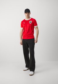Crvena nogometna majica s bijelim naglascima, koja sadrži nacionalni grb i logotip. Uparena s crnim hlačama i bijelim tenisicama. Minimalistička pozadina.