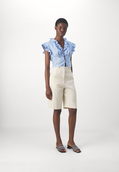 Blouse à rayures bleu et blanc à manches courtes avec volants, associée à un short beige bien taillé et des sandales à enfiler noires avec des accents décoratifs sur les brides.
