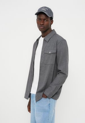 Mann trägt eine graue, geknöpfte Hemdjacke, weißes T-Shirt, hellblaue Jeans und eine graue Baseballkappe und steht vor einem weißen Hintergrund.