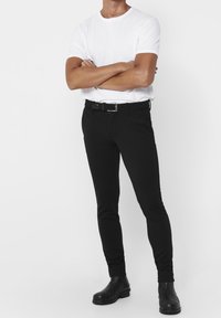 Schwarze Slim-Fit-Hose aus elastischem Stoff, mit einem gewebten Gürtel, kombiniert mit einem schlichten weißen T-Shirt mit Rundhalsausschnitt und schwarzen Stiefeln.