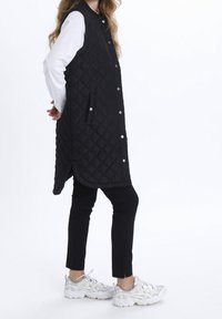 Persona con chaleco largo acolchado negro sobre camisa blanca de manga larga, pantalones negros y zapatillas deportivas blancas, de pie de lado con las manos detrás de la espalda.