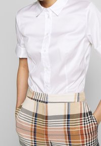 Witte korte mouwen blouse met knoopsluiting aan de voorkant, gecombineerd met beige en geruite hoge taillebroek met oranje, blauwe en zwarte accenten.