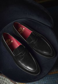 Zwarte leren instappers met een slank ontwerp, voorzien van een zadeltas, rode binnenzolen en gedetailleerde stiksels langs de randen.